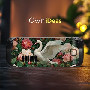 May include: A black handheld gaming device with a floral design featuring a white swan and pink peonies. The device has the text "OwniIdeas" in white. The background is a blurred mix of green and brown.