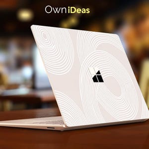 Könnte beinhalten: Ein Laptop mit einer weißen Schutzfolie, die ein Muster aus konzentrischen kreisförmigen Linien und ein schwarzes Microsoft-Logo aufweist. Der Text OwniDeas ist oben sichtbar. Der Laptop ist von hinten zu sehen und zeigt das Deckeldesign.