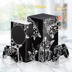 Puede incluir: Consola de juegos y mandos negros con un diseño de boceto de gato blanco. La consola y los mandos tienen un patrón repetido de caras de gato. El texto "OwniDeas" está en la esquina superior izquierda.