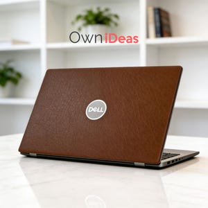 K&ouml;nnte beinhalten: Ein brauner Laptop-Skin mit Lederstruktur und Dell-Logo. Der Laptop ist ge&ouml;ffnet und zeigt Tastatur und Bildschirm. Der Hintergrund umfasst ein wei&szlig;es Regal mit Pflanzen und den Text "OwniIdeas".