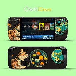 May include: A black handheld gaming console with a colourful cat and space-themed design. The console has a screen displaying the game "Cocoon" and the text "Play Aperture Desk Job".