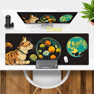 以下が含まれることがあります： 黒と白のデスクマットに、猫と惑星のカラフルなイラストが描かれています。猫はオレンジと白で、尻尾を体の周りに巻き付けています。惑星は猫の周りを円形に配置されており、黄色、オレンジ、青、緑の色合いで描かれています。デスクマットは木製の表面に置かれています。マットには「OwnIdeas」という文字が印刷されています。