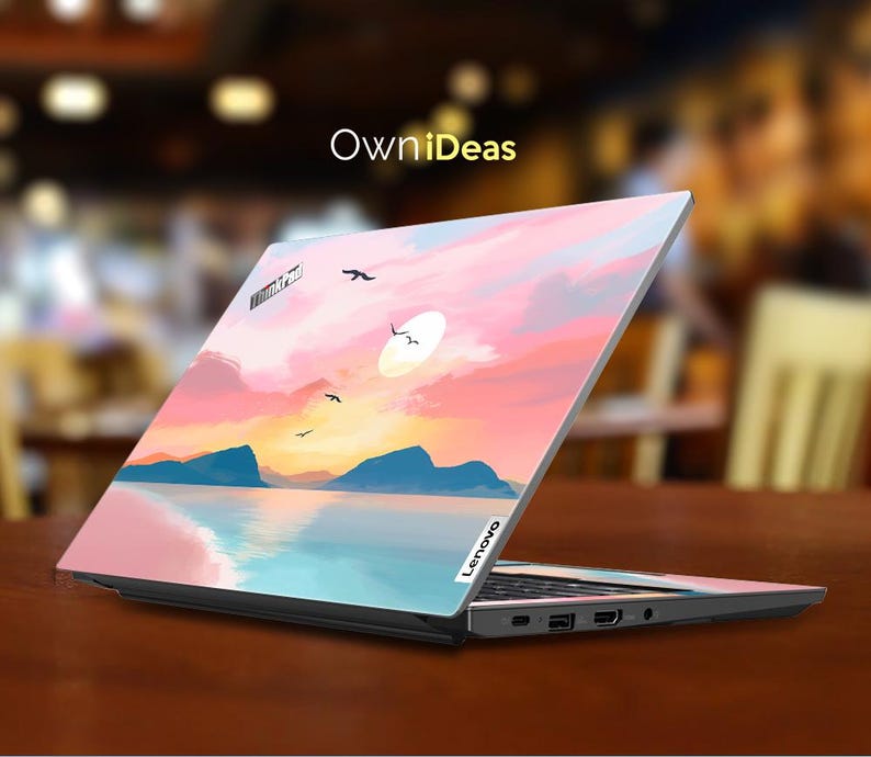 Op de afbeelding: Lenovo laptop met een aangepaste skin met een zonsondergang boven water. Het ontwerp bevat roze, oranje en blauwe tinten, met bergen en vogels in silhouet. De laptop staat open op een bruin oppervlak.