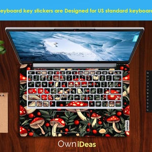 May include: Laptop with a keyboard skin featuring red and white mushrooms, green leaves, and red berries on a black background. The keyboard keys are black and white. The screen displays a landscape. Text at the top says, "Keyboard key stickers are Designed for US standard keyboards."
