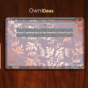 Op de afbeelding: Een laptop skin met een bloemenpatroon, met een lavendelkleurige achtergrond en perzikkleurige bladeren. De skin bedekt de onderkant van de laptop, met de tekst "OwniDeas". Een klein notitieboekje en een zwart notitieboekje zijn zichtbaar.
