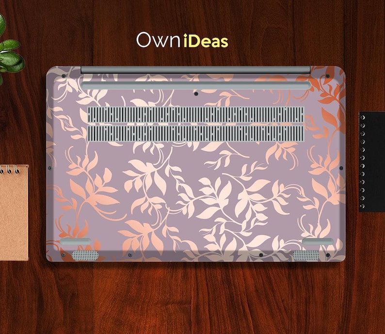 Op de afbeelding: Een laptop skin met een bloemenpatroon, met een lavendelkleurige achtergrond en perzikkleurige bladeren. De skin bedekt de onderkant van de laptop, met de tekst "OwniDeas". Een klein notitieboekje en een zwart notitieboekje zijn zichtbaar.