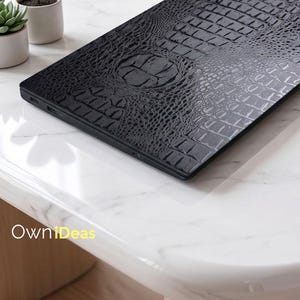 May include: A black laptop with a textured, crocodile-skin pattern on the lid. The laptop is resting on a white marble surface. The design includes a circular emblem in the center of the lid.
