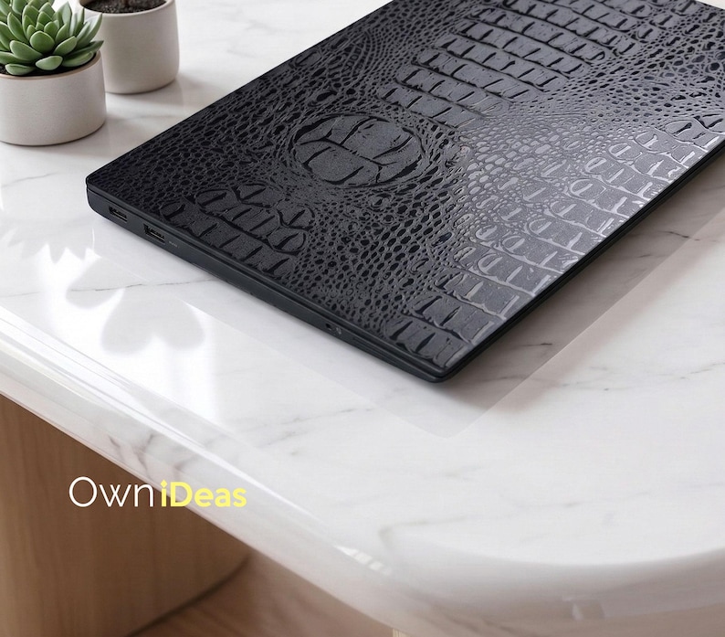 May include: A black laptop with a textured, crocodile-skin pattern on the lid. The laptop is resting on a white marble surface. The design includes a circular emblem in the center of the lid.