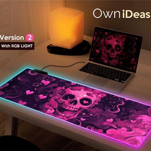 Op de afbeelding: Een roze en paarse gaming muismat met een schedel- en hartendesign. De muismat heeft een oplichtende roze en blauwe rand. De tekst "OwnIdeas" en "Version 2 with RGB LIGHT" is zichtbaar in de afbeelding.