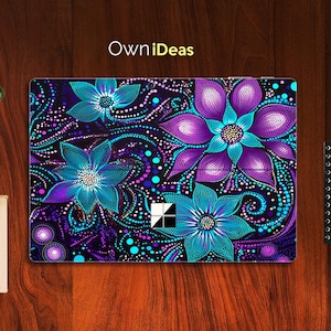 Puede incluir: Una piel para portátil con un diseño abstracto colorido con flores moradas, azules y turquesas sobre un fondo negro. El diseño está formado por pequeños puntos y remolinos. La piel es compatible con dispositivos Microsoft Surface.