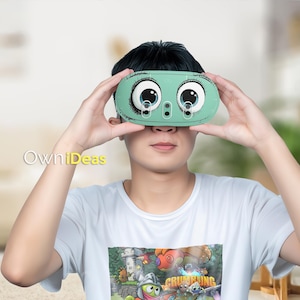 May include: A mint green VR headset with cartoon eyes. The headset is held up to the face. The text "OwniDeas" is in the lower left corner. The person is wearing a white t-shirt with a colourful graphic.