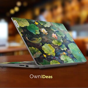 Puede incluir: Un ordenador port&aacute;til con un dise&ntilde;o floral en la tapa. El dise&ntilde;o presenta nen&uacute;fares, hojas y una rana. El port&aacute;til est&aacute; abierto y la pantalla est&aacute; apagada. El texto "Lenovo" es visible en la tapa.