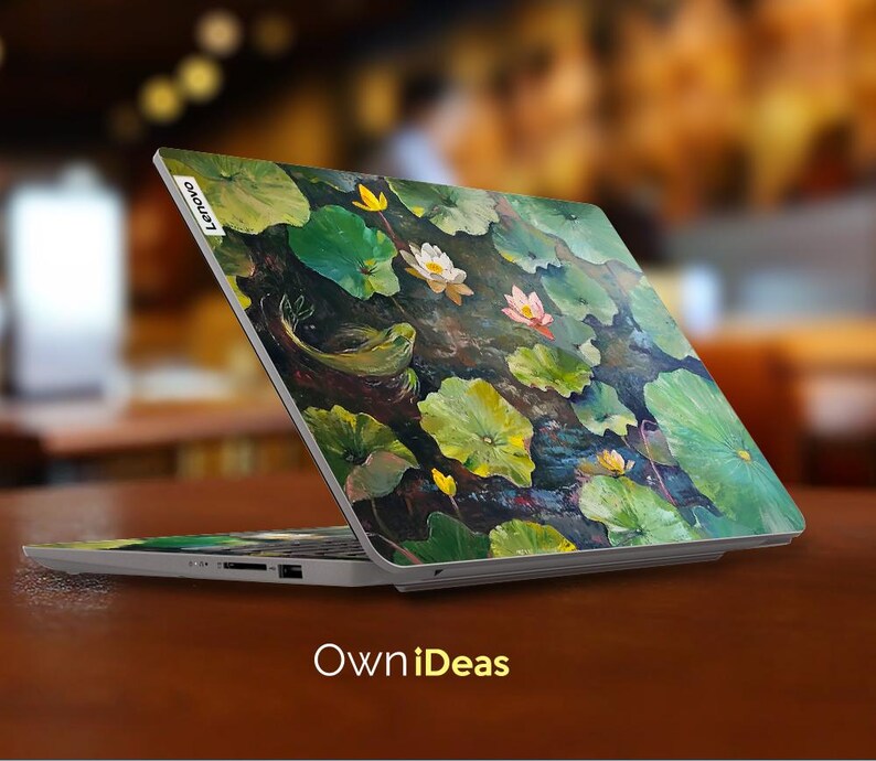 Puede incluir: Un ordenador port&aacute;til con un dise&ntilde;o floral en la tapa. El dise&ntilde;o presenta nen&uacute;fares, hojas y una rana. El port&aacute;til est&aacute; abierto y la pantalla est&aacute; apagada. El texto "Lenovo" es visible en la tapa.