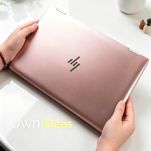 May include: A rose gold HP laptop with the HP logo in the center. The laptop is open and held by two hands. The laptop is on a white surface. The text "OwniDeas" is visible in the bottom left corner.