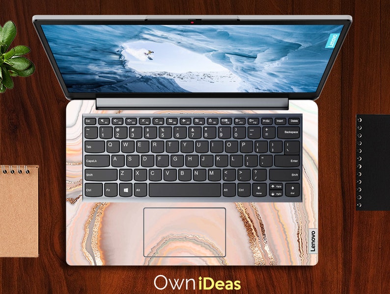 Op de afbeelding: Een laptop met een wit en gouden marmerontwerp op de deksel en het toetsenbord. De laptop is gesloten en het scherm toont een besneeuwde berg sc&egrave;ne. De tekst "Own iDeas" is onderaan de afbeelding gedrukt.