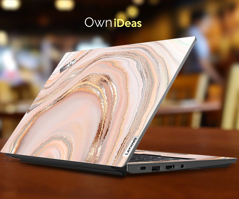 Op de afbeelding: Een laptop met een roze en gouden marmerontwerp op de deksel. De laptop is gesloten en de deksel is open om het ontwerp te laten zien. De laptop staat op een houten tafel.
