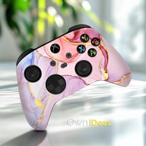Puede incluir: Un mando de Xbox con un patrón de mármol rosa, morado y dorado. El mando tiene un fondo blanco con remolinos rosas, morados y dorados. Los botones son negros y el logotipo de Xbox es blanco.