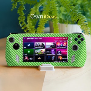 May include: A handheld gaming device with a green carbon fiber textured skin. The screen displays a game library with various game icons. The device is on a wooden surface with a fern in the background. The text "OwniDeas" is visible.