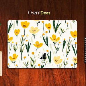 Puede incluir: Una piel de portátil blanca con un patrón floral con flores amarillas y hojas verdes. La piel tiene el texto "OwnIdeas" en la parte superior.
