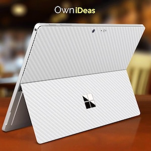 May include: White laptop computer with a carbon fibre pattern skin. The laptop is closed and the screen is facing away from the camera. The laptop has a Microsoft logo on the lid.