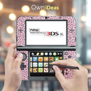 May include: A pink and black leopard print Nintendo 3DS XL handheld game console. The screen displays the words "new Nintendo 3DS XL". The console is held in two hands, with a stylus touching the touchscreen. The brand name "OwniIdeas" is in the top left corner.