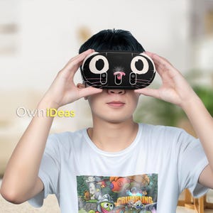 May include: A black VR headset with a cat face design, held up to the eyes. The headset has large white eyes, pink nose, and whiskers. The person is wearing a white t-shirt with a colorful graphic.