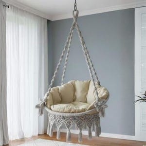 May include: A hanging macrame chair featuring intricate off-white knotted rope patterns, a circular base with decorative fringe, and thick, plush beige cushions. The chair is suspended by sturdy braided ropes that converge at a single top point.