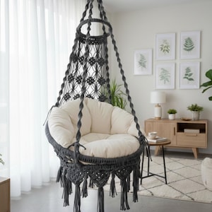 Op de afbeelding: Een hangende macramé stoel met een crèmekleurig kussen en donkergrijze macramé details. De stoel hangt aan het plafond en heeft decoratieve kwastjes. De stoel staat in een woonkamer.