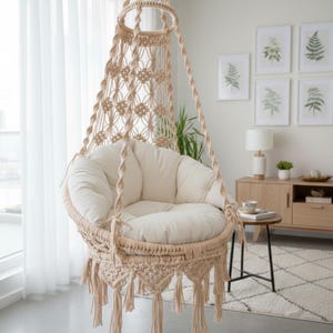 Puede incluir: Una silla colgante de macramé beige con un cojín color crema. La silla está suspendida de un anillo circular y presenta intrincados patrones tejidos y borlas decorativas. La silla está diseñada para uso en interiores y añade un toque bohemio.