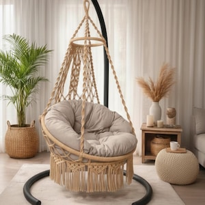 Puede incluir: Una silla colgante de macramé beige con un asiento redondo y acolchado. La silla está suspendida de un marco de metal negro y presenta flecos en la parte inferior. Una planta en maceta y objetos decorativos están en el fondo.