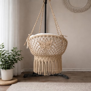 Könnte beinhalten: Eine beige Makramee-Babywiege mit Schmetterlingsdesign. Die Wiege ist an einem schwarzen Metallständer aufgehängt. Die Wiege hat eine weiße Innenausstattung und eine Fransen am Boden. Der Hintergrund umfasst eine Pflanze in einem weißen Topf und einen Teppich.