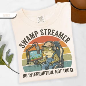 May include: Ivory-colored t-shirt featuring a cartoon frog gamer with the text "SWAMP STREAMER" above and "NO INTERRUPTION. NOT TODAY." below. The frog is wearing a hat and headphones, sitting in a gaming chair.