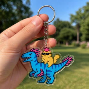 May include: A clear acrylic keychain featuring a blue dinosaur with pink accents and a yellow cat wearing sunglasses and a crown. The keychain has a silver keyring and chain.