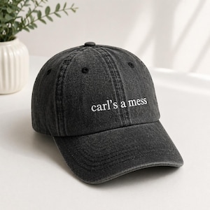 Puede incluir: Gorra de béisbol gris oscuro con visera curva. La gorra presenta el texto bordado "carl's a mess" en blanco. La gorra tiene un botón en la parte superior y está hecha de tela texturizada.