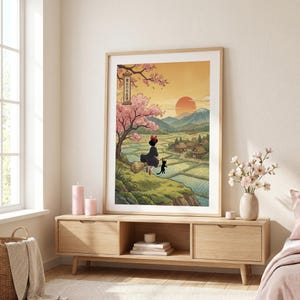 May include: A framed art print featuring a Japanese-inspired illustration. A young witch with a black cat sits on a broom, overlooking a village and rice fields. The artwork includes a cherry blossom tree, a setting sun, and Japanese text.