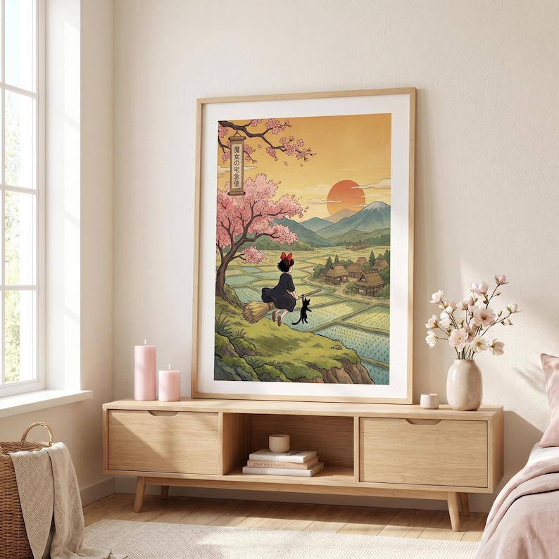 May include: A framed art print featuring a Japanese-inspired illustration. A young witch with a black cat sits on a broom, overlooking a village and rice fields. The artwork includes a cherry blossom tree, a setting sun, and Japanese text.