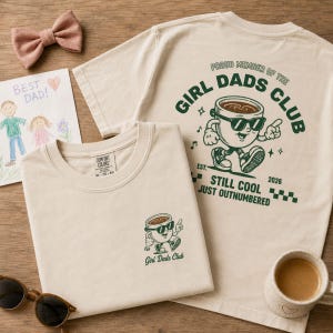 Pode incluir: Uma t-shirt bege com um pequeno gráfico na frente e um gráfico grande nas costas. O design das costas mostra uma chávena de café de desenho animado com óculos de sol e o texto "PROUD MEMBER OF THE GIRL DADS CLUB" e "STILL COOL JUST OUTNUMBERED".