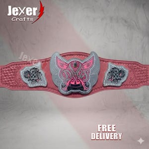 May include: A pink wrestling championship belt with a central emblem featuring a pink and silver design. The belt has two side plates with a silver and black design. The text "Jexer Crafts" is visible in the top left corner.