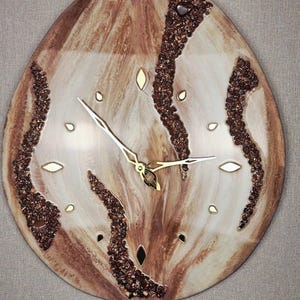 May include: A teardrop-shaped wall clock featuring a marbled brown and cream resin surface. The clock face is accented with three vertical, textured bands of dark brown crushed stone and small, gold-toned diamond-shaped hour markers.
