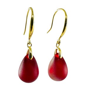 May include: A pair of gold-colored drop earrings with teardrop-shaped, deep red glass pendants. The earrings have a simple, elegant design, with the red glass contrasting against the gold hooks.