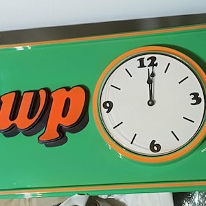 May include: A rectangular green wall clock featuring the orange text Newp ort with an integrated circular clock face in the center. The clock face has a white background with black numerals and hands. A small white rectangular label displays text.