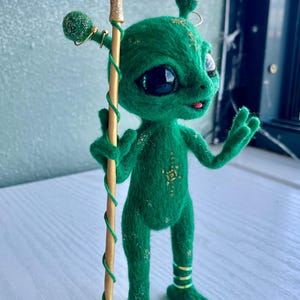 Könnte beinhalten: Eine grüne, nadelgefilzte Alien-Figur mit großen schwarzen Augen, die einen goldenen Stab mit einer Mondsichel, einem klaren Kristall und grünen Sternen hält. Die Figur hat goldene Glitzerdetails am Körper und grüne Bänder an den Beinen.