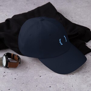 May include: A navy blue baseball cap with a curved brim. The cap features a light blue embroidered design of curly braces. A black jacket and a wristwatch with a brown leather band are in the background.