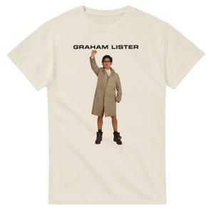 Könnte beinhalten: Ein cremefarbenes T-Shirt mit einer Grafik einer Person in einem beigen Trenchcoat, dunkler Brille und dunklen Stiefeln, wobei ein Arm zur Faust erhoben ist. Der Text GRAHAM LISTER ist in schwarzen Großbuchstaben über der Figur aufgedruckt.