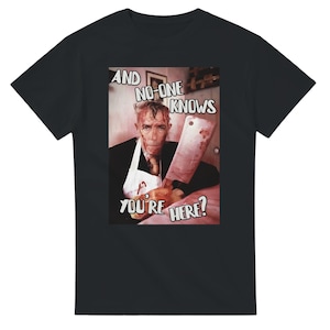 May include: Black t-shirt with a graphic image of a man holding a bloody cleaver. The image includes the text "AND NO-ONE KNOWS YOU'RE HERE?" in a white, stylized font. The man is wearing a white apron and a tie.