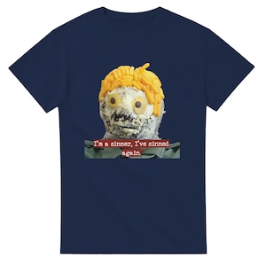 May include: A navy blue t-shirt with a food-based face design in yellow, orange, and white. The text "I'm a sinner, I've sinned again" is printed in red below the image. A unique and quirky design.