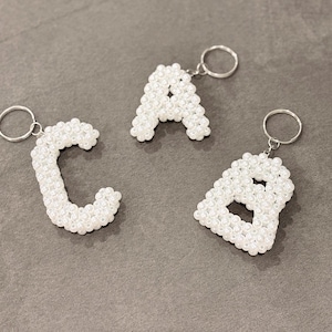 Peut inclure: Trois porte-clés en forme de lettres, perlés de blanc, épelant les lettres "C", "A" et "B". Chaque lettre est faite de petites perles blanches rondes et attachée à un porte-clés argenté. Les porte-clés sont présentés sur une surface grise.