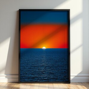 May include: A framed vertical wall art print featuring a vibrant sunset over a dark blue ocean. The horizon line separates a deep orange and red sky from the textured, rippling water below, with a small white sun centred at the horizon line.