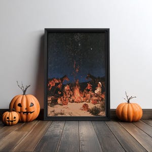 May include: A framed rectangular print depicting a nighttime scene of cowboys gathered around a campfire under a starry sky, flanked by two orange jack-o'-lanterns on the left and one orange pumpkin on the right on a wooden floor.