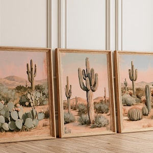 May include: Three framed vertical art prints depicting desert landscapes with saguaro and prickly pear cacti. The illustrations feature a soft color palette of muted greens, dusty pinks, and sandy tans, showcasing arid terrain under a pale sky.